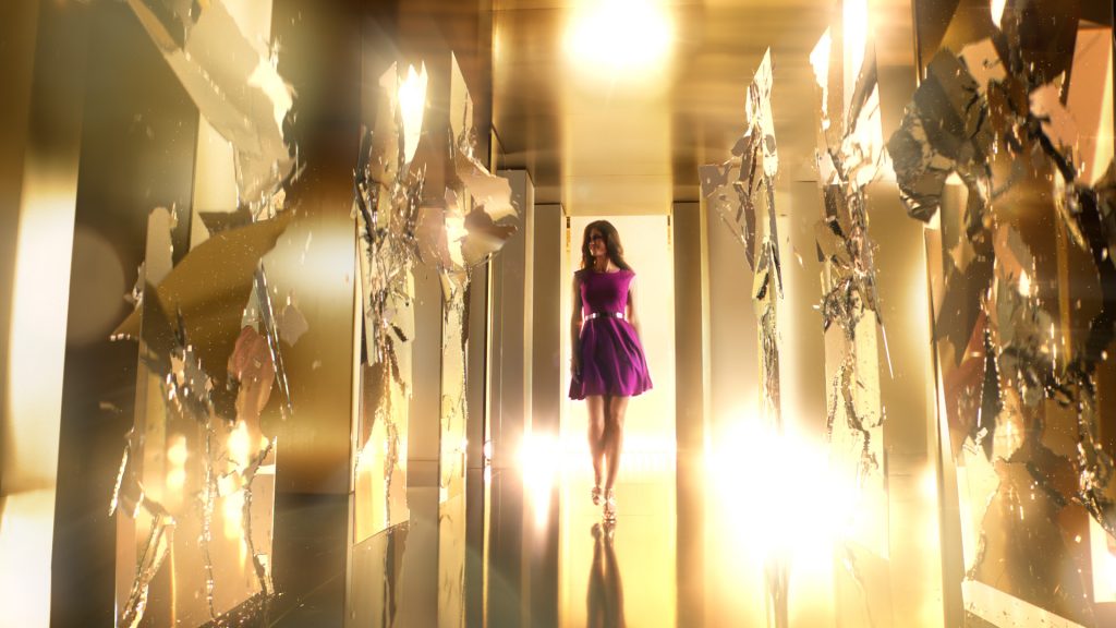 A woman in a purple wool dress walking through a bright golden VFX rendered room with lots of shattering mirrors.