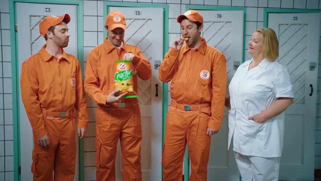 Three men in orange overalls standing beside a stout, blonde nurse in white uniform, enjoying some Zweifel "Cractiv" potato-chips.